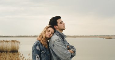 woman in blue denim jacket leaning on man's shoulder near body of water