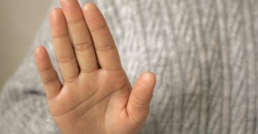 Crop anonymous African American female with curly hair in sweater showing stop gesture against light wall in room
