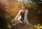 Woman in tranquil meditation pose outdoors, bathed in sunlight, surrounded by lush greenery.