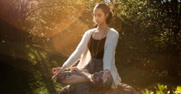 Woman in tranquil meditation pose outdoors, bathed in sunlight, surrounded by lush greenery.